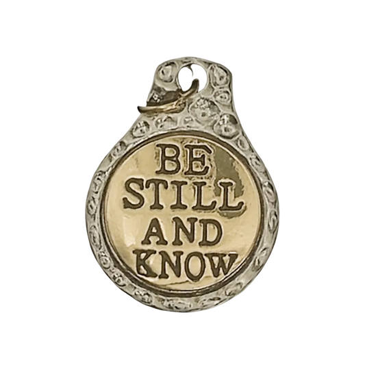 close-up of round two-tone charm reading BE STILL AND KNOW on black background