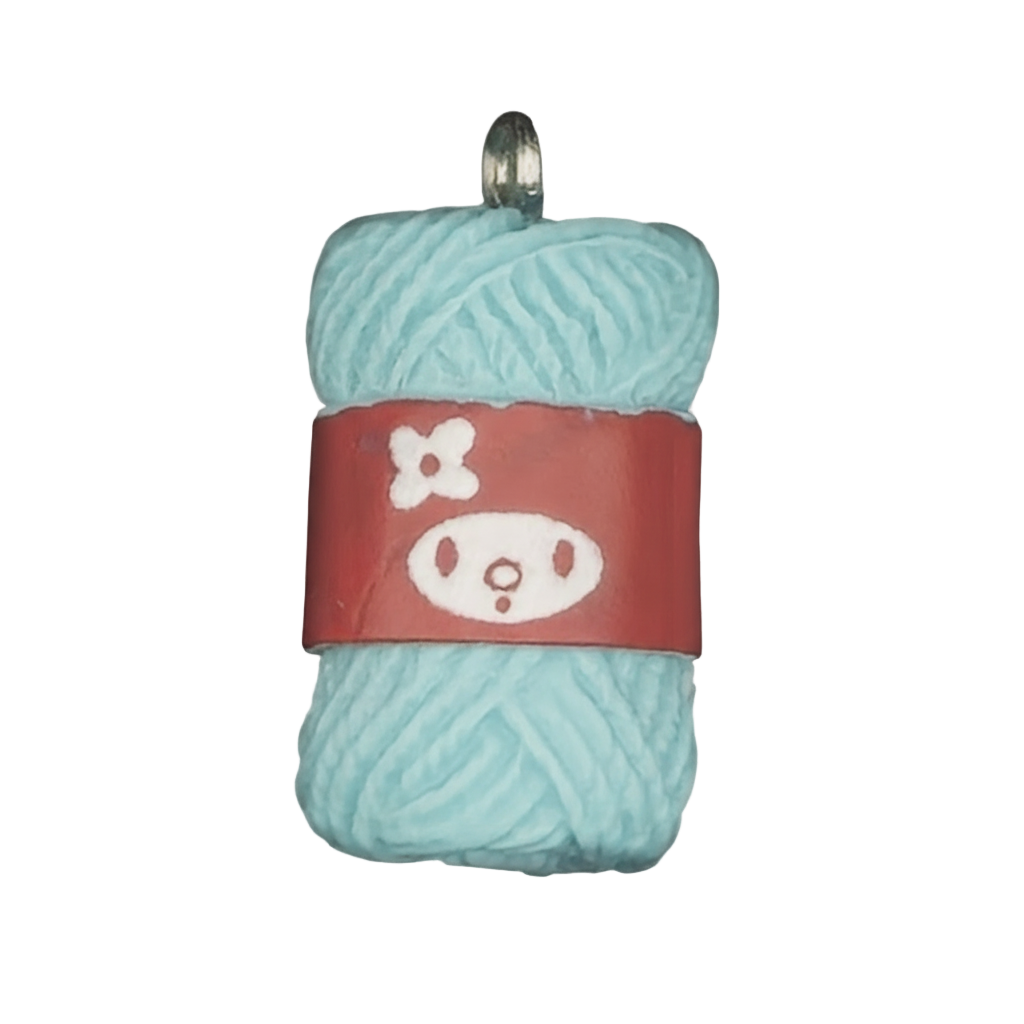 Close-up of a light blue yarn skein charm with a red label showing a simple face and flower on a black background.