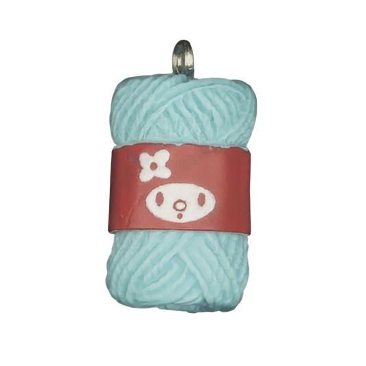 Close-up of a light blue yarn skein charm with a red label showing a simple face and flower on a black background.