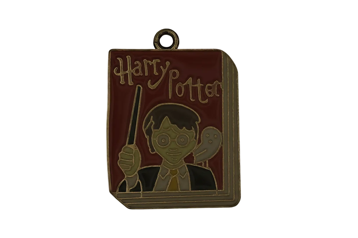 Close-up of an enamel, book-shaped charm with 'Harry Potter' text, a glasses-wearing boy holding a wand, and an owl on a red cover with gold outline.