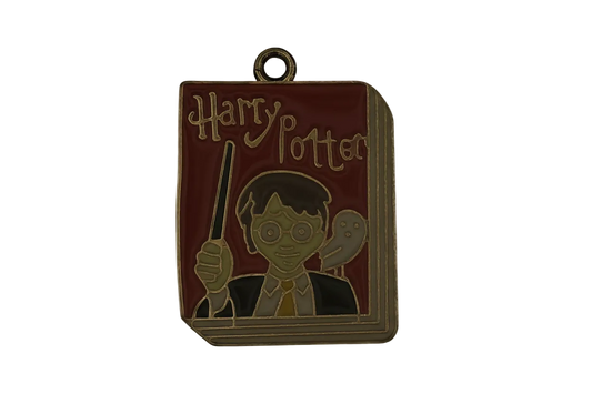 Close-up of an enamel, book-shaped charm with 'Harry Potter' text, a glasses-wearing boy holding a wand, and an owl on a red cover with gold outline.